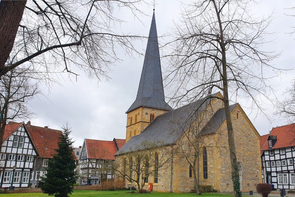 Die gelbe Sandsteinfassade der Apostelkirche in Gütersloh mit ihrem markanten, spitzen Turmhelm inmitten von Bäumen.