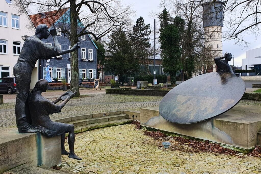 Der Rondellbrunnen in Gütersloh mit Bronzeskulpturen und einer schrägen Metallscheibe, im Hintergrund ragt der historische Wasserturm empor.
