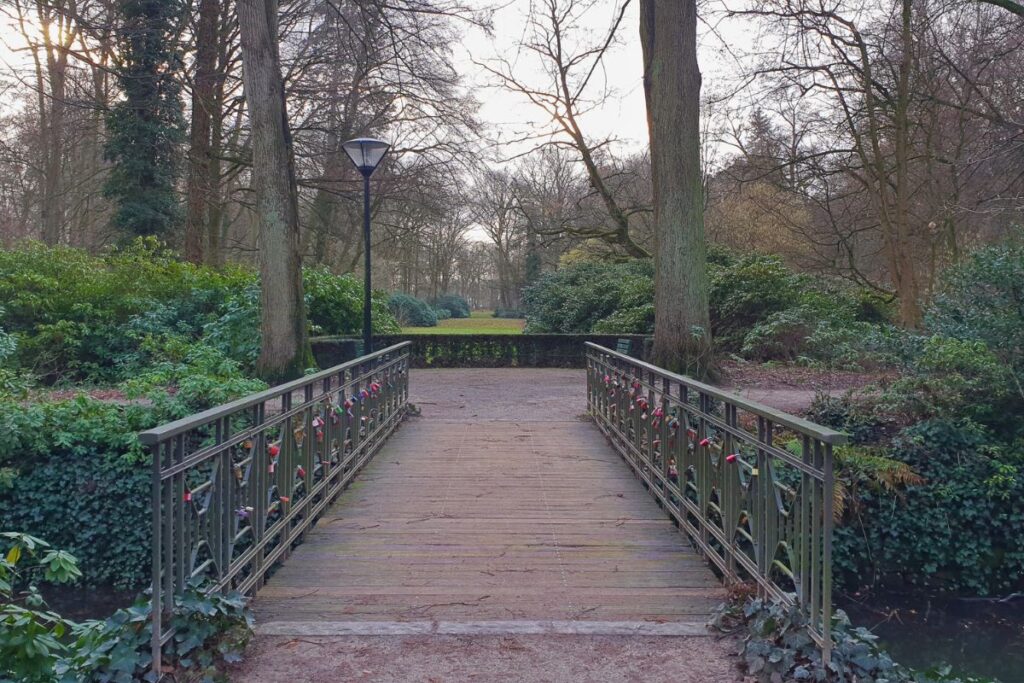 Eine hölzerne Brücke mit bunten Liebesschlössern im Gütersloher Stadtpark, umgeben von hohen, dichten Bäumen.
