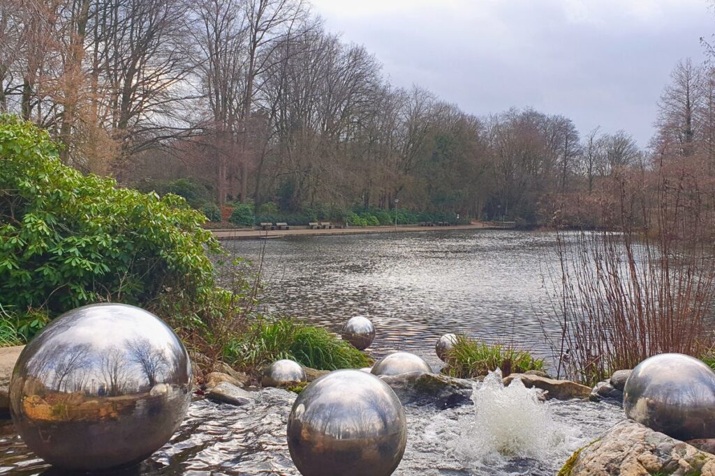 Große, spiegelnde Metallkugeln liegen in einem kleinen Wasserlauf am Ibrüggers Teich im Stadtpark Gütersloh.