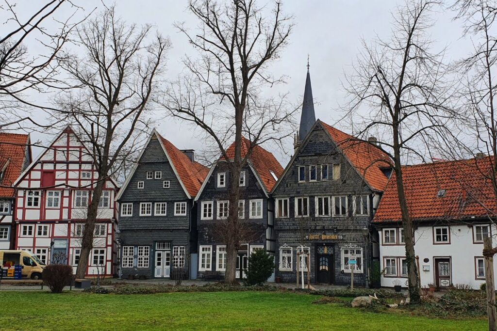 Blick über eine grüne Wiese auf die historische Häuserzeile am Alten Kirchplatz in Gütersloh mit der Spitze des Kirchturms im Hintergrund.
