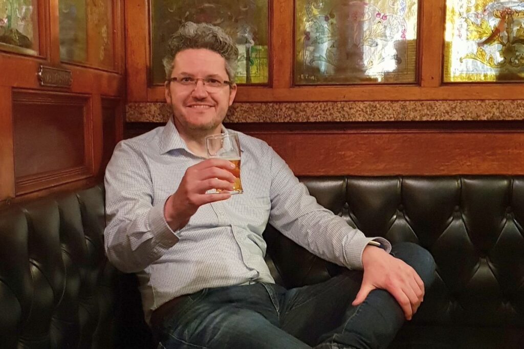 Reiseblogger Nicolo Martin sitzt lächelnd mit einem Glas Bier in einer gemütlichen Lederecke eines traditionellen irischen Pubs mit buntem Glasfenster.