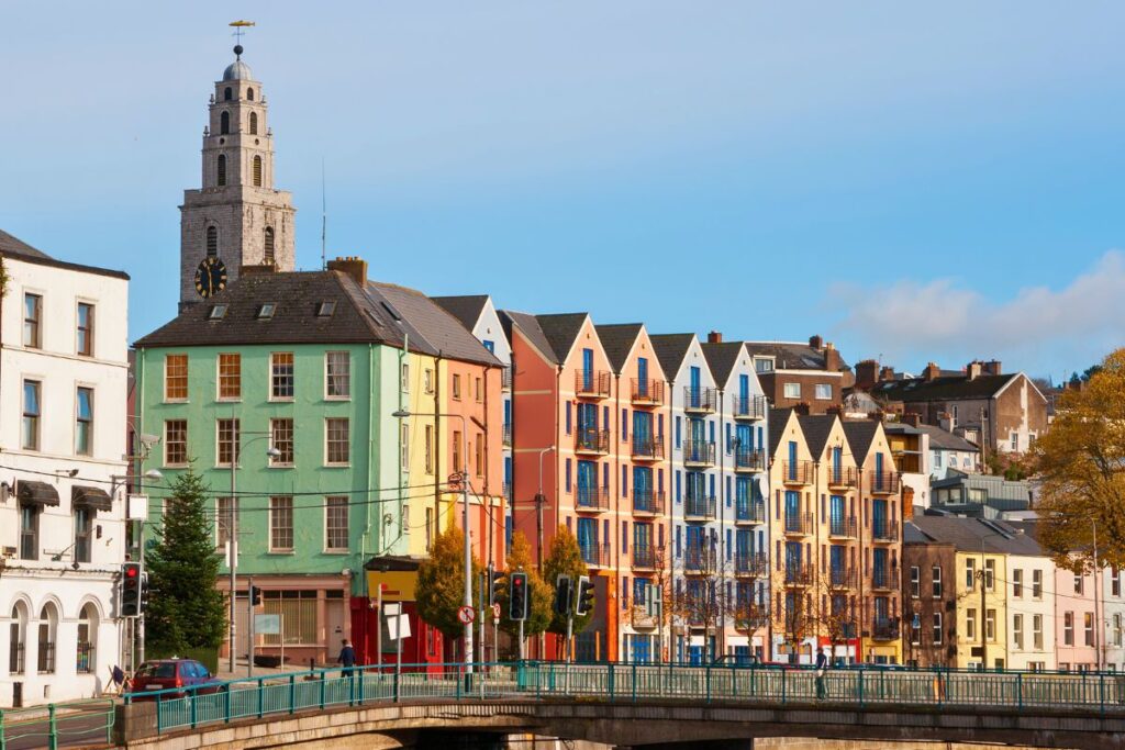 Eine Reihe farbenfroher Häuser am Flussufer in Cork, im Hintergrund ragt der Shandon Tower unter blauem Himmel empor.