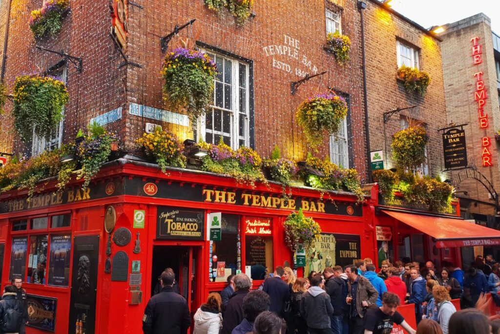 Die ikonische rote Fassade des „The Temple Bar“ Pubs in Dublin, üppig geschmückt mit Blumenampeln, umgeben von feiernden Menschenmengen.