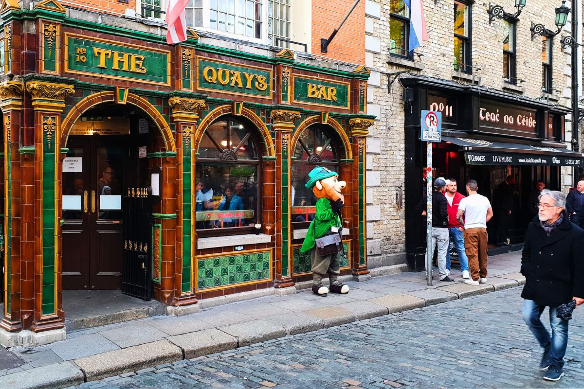 Die kunstvoll verzierte, grün-goldene Fassade der Quays Bar in Dublin, davor steht ein Leprechaun-Maskottchen auf einer belebten Kopfsteinpflasterstraße.