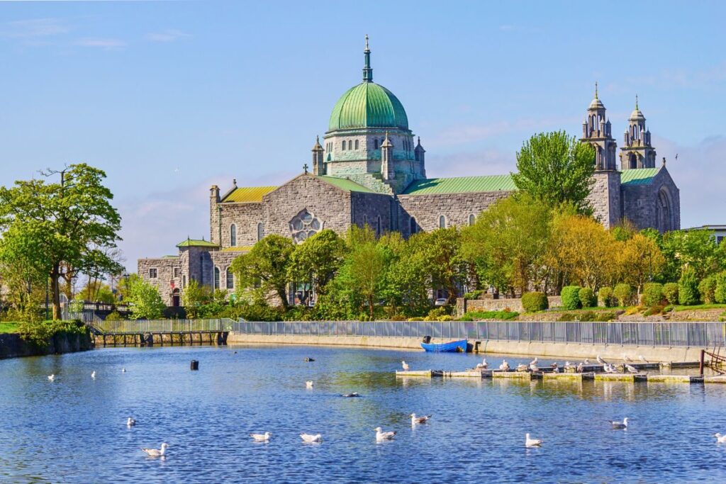 Die imposante Galway Cathedral mit ihrer markanten grünen Kuppel, umgeben von üppigem Grün am Ufer des Corrib-Flusses mit Schwänen im Wasser.