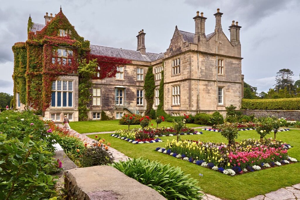 Das herrschaftliche Muckross House in Killarney mit efeubewachsenen Steinmauern und einem akkurat gepflegten, bunten Blumengarten im Vordergrund.