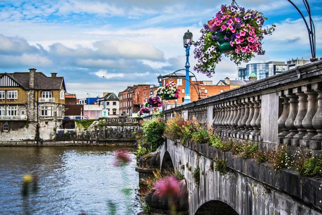 Blick von einer mit Blumenampeln geschmückten Steinbrücke auf den Shannon in Limerick mit historischen Gebäuden am Ufer.