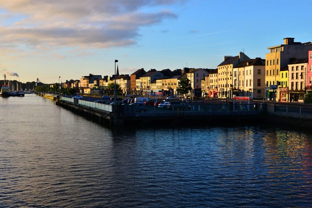 lick über das glitzernde Wasser des Flusses Suir auf die Uferpromenade von Waterford während eines goldenen Sonnenuntergangs mit historischen Gebäuden am Kai.