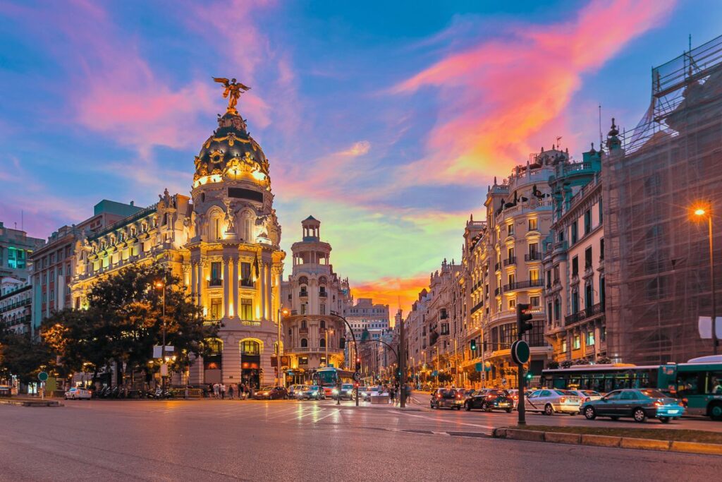 Die beleuchtete Gran Vía in Madrid mit dem ikonischen Metropolis-Gebäude unter einem dramatisch pinken Abendhimmel.