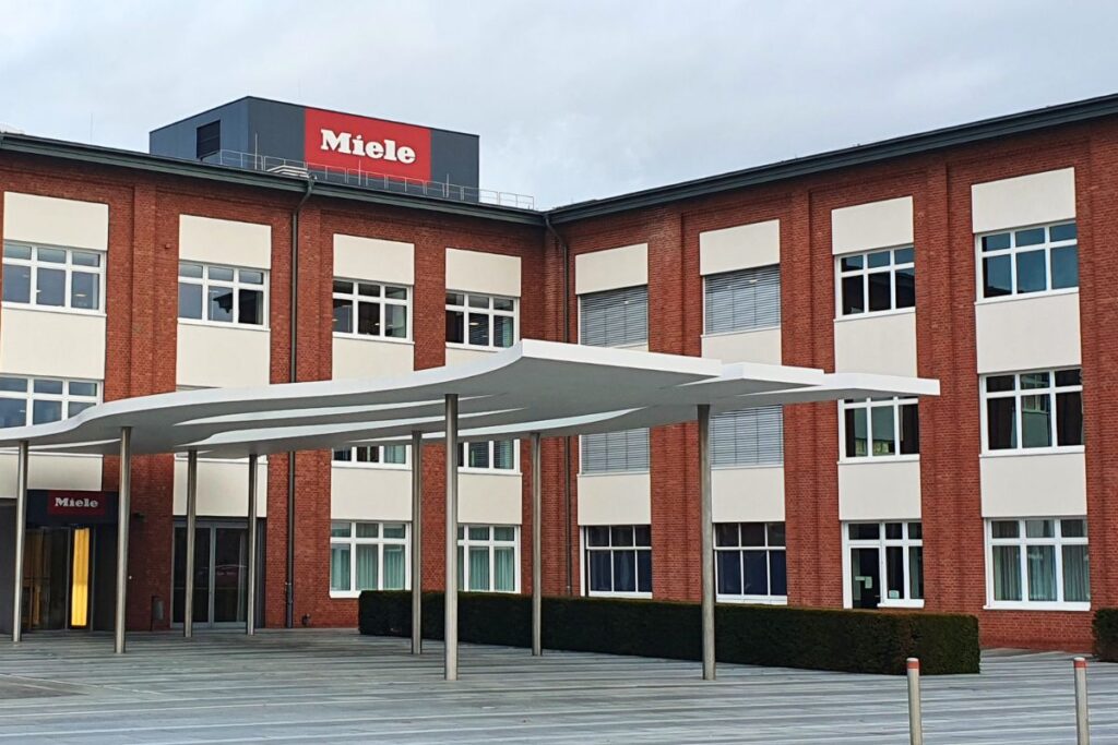Das Miele Museum in Gütersloh, ein Backsteingebäude mit modernem weißem Vordach und dem roten Miele-Logo an der Fassade.