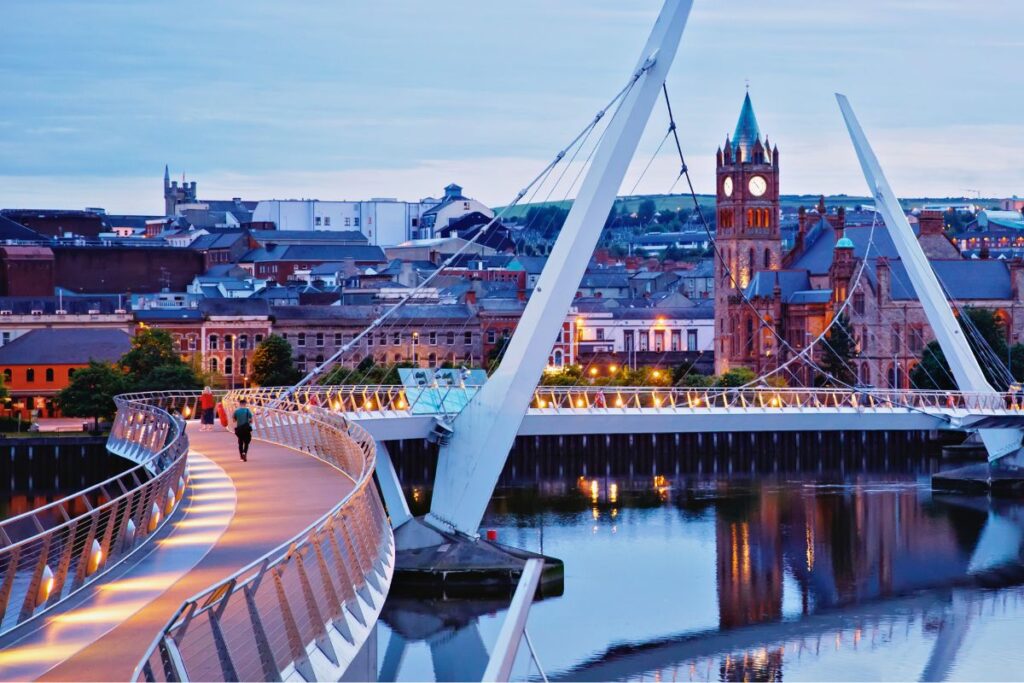 Die beleuchtete Peace Bridge in Derry, die sich abends geschwungen über den Fluss Foyle erstreckt, mit historischen Gebäuden im Hintergrund.