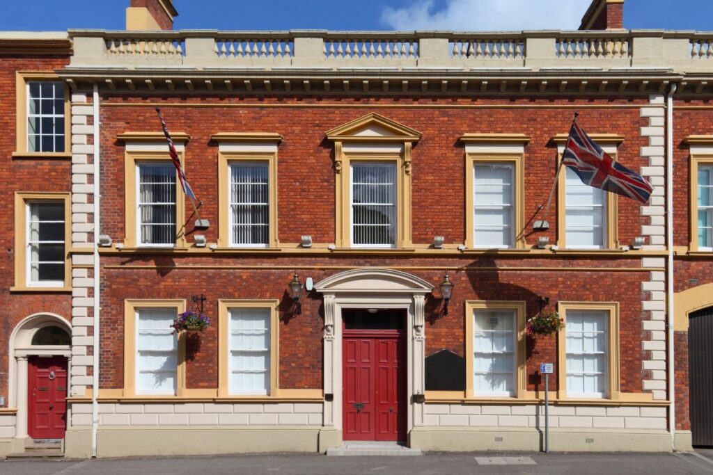 Die symmetrische rote Backsteinfassade eines historischen Gebäudes in Lisburn, Nordirland, mit wehenden Union Jack Flaggen unter blauem Himmel.