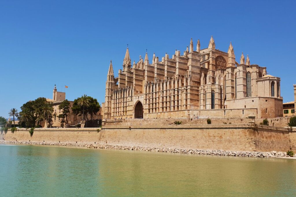 Die imposante sandsteinfarbene Kathedrale La Seu in Palma de Mallorca spiegelt sich im Wasserbecken des Parc de la Mar unter strahlend blauem Himmel.