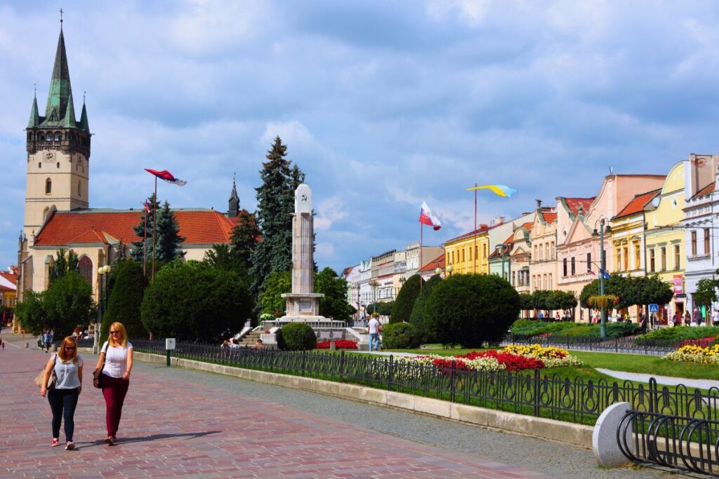 Ein weiter Blick über den Hauptplatz (Hlavná ulica) in Prešov mit der St.-Nikolaus-Konkathedrale, Blumenbeeten und wehenden Flaggen.