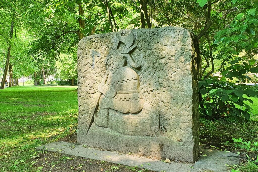 Steinskulptur des „Bornschissers“ in einer grünen Parkanlage in Rotenburg an der Fulda, umgeben von schattigen Bäumen.