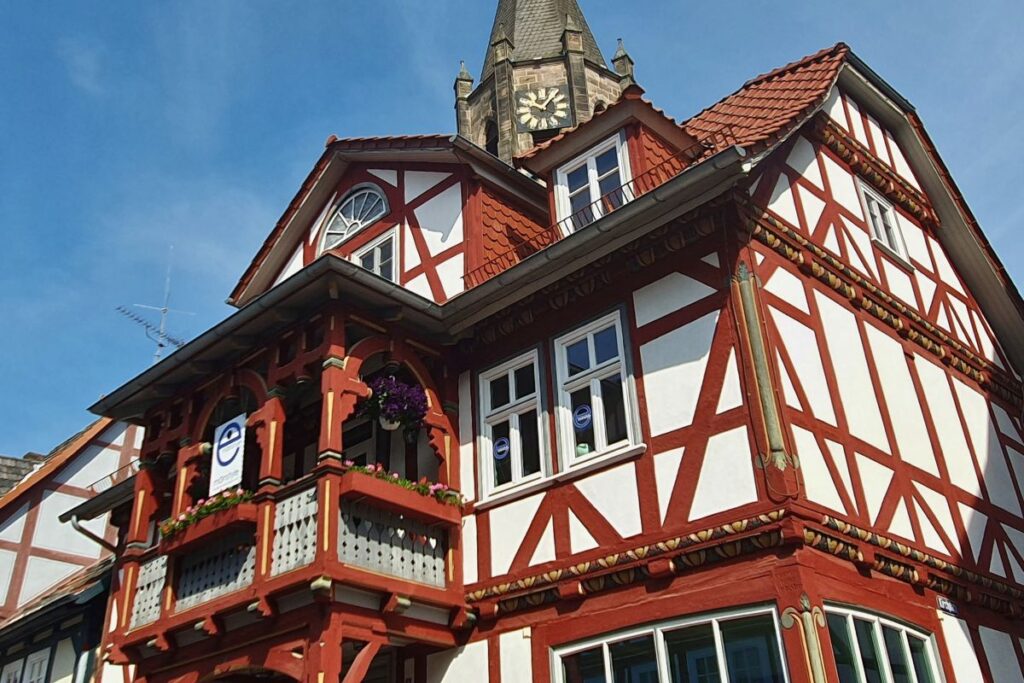 Ein wunderschön restauriertes Fachwerkhaus, das Alte Schulhaus in Rotenburg an der Fulda, mit roten Balken und Blumen auf dem Balkon vor einem blauen Himmel.