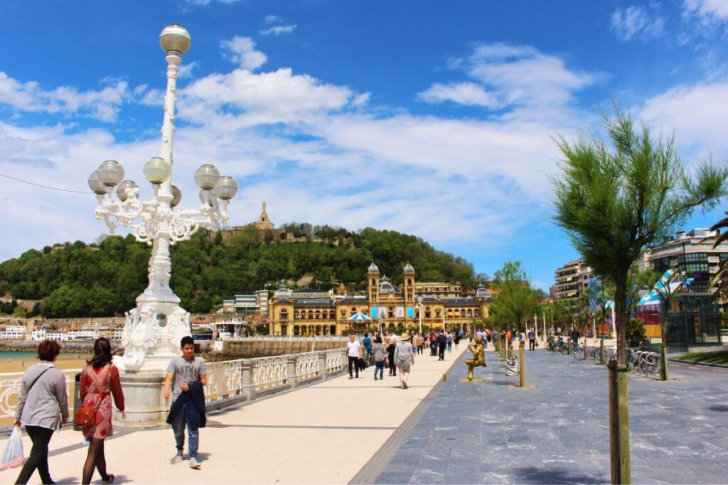Spaziergänger auf der eleganten weißen Uferpromenade La Concha in San Sebastian mit den markanten weißen Laternen und Blick auf die bewaldeten Hügel.