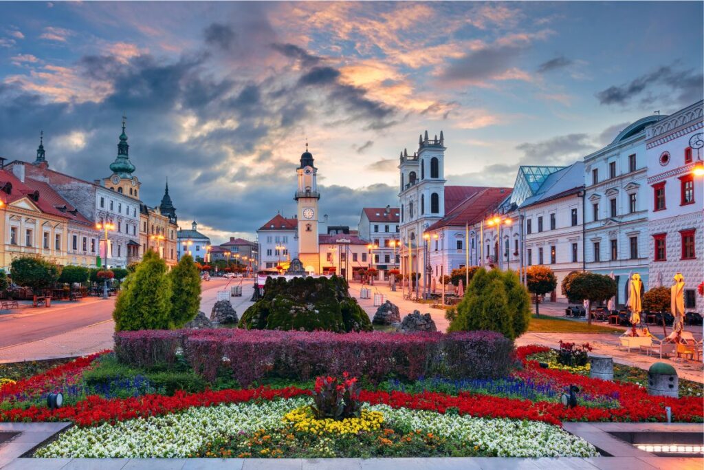 Der weite SNP-Platz in Banská Bystrica mit dem historischen Uhrturm, bunten Blumenbeeten und Bergen im Hintergrund bei Sonnenuntergang.