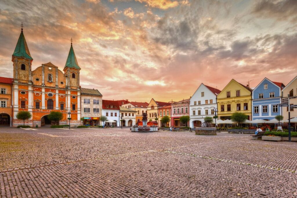 Eine Reihe farbenfroher Bürgerhäuser mit Arkaden am Mariánske-Platz in Žilina vor einem dramatischen, orangefarbenen Abendhimmel.