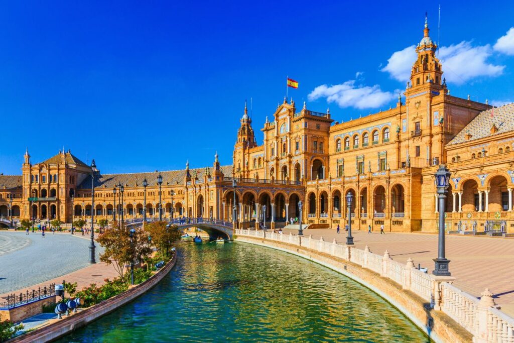 Die beeindruckende Plaza de España in Sevilla mit ihrem halbrunden Gebäude, dem Kanal mit Booten und den verzierten Brücken unter tiefblauem Himmel.