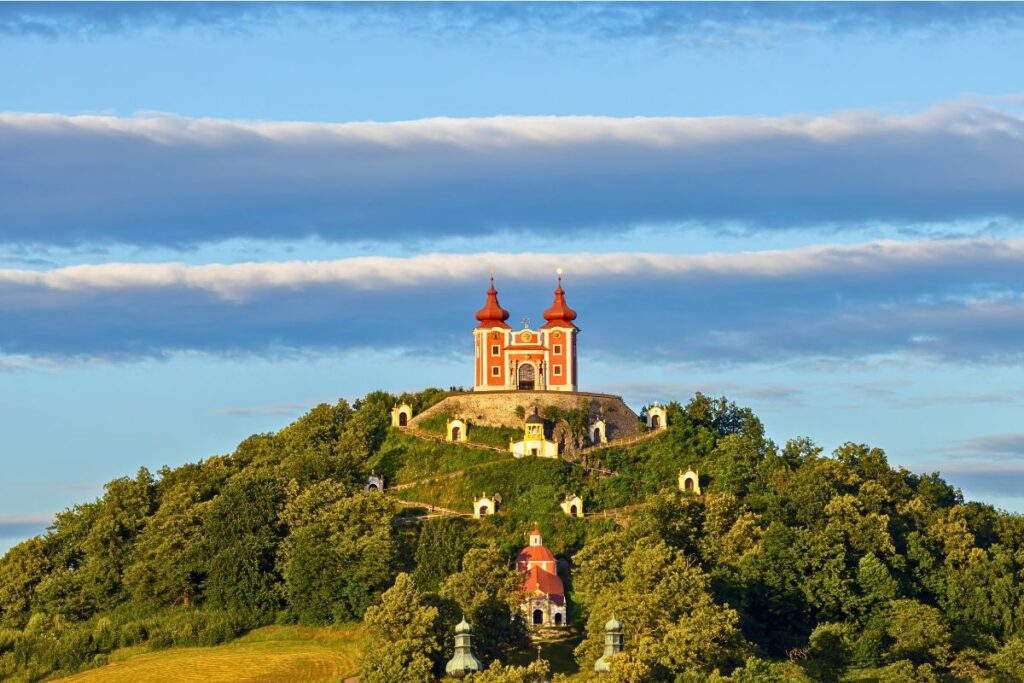 Die rot-weißen Kirchengebäude des Kalvarienbergs (Kalvária) auf einem grünen Hügel in Banská Štiavnica unter einem dramatischen Wolkenhimmel.