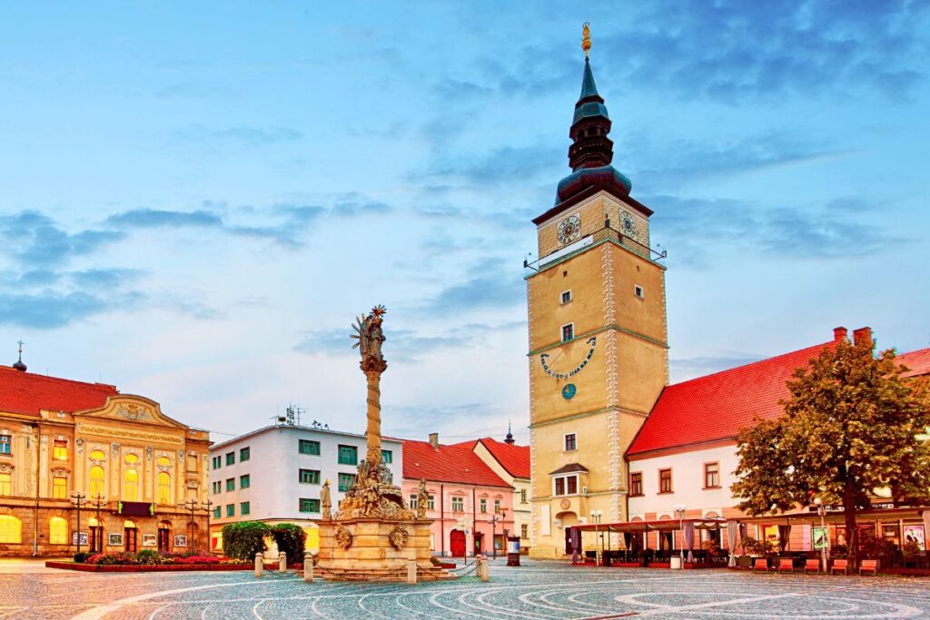 Der weitläufige Trojičné-Platz in Trnava mit dem beleuchteten Stadtturm, dem Rathaus und der Dreifaltigkeitssäule bei Dämmerung.