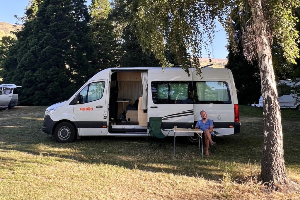 Nicolo Martin sitzt entspannt an einem Campingtisch vor seinem weißen Apollo-Camper-Van im Schatten von Bäumen.