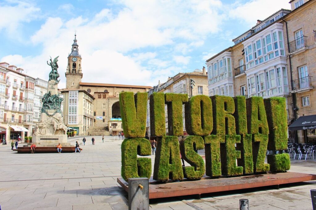 Die markante grüne Moos-Skulptur „VITORIA GASTEIZ!“ auf der Plaza de la Virgen Blanca vor historischen Denkmälern und Gebäuden.