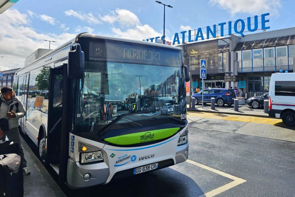 Ein weiß-grüner „Naolib“-Flughafenbus am Terminal des Flughafens Nantes Atlantique, im Hintergrund die blaue Aufschrift des Flughafennamens.