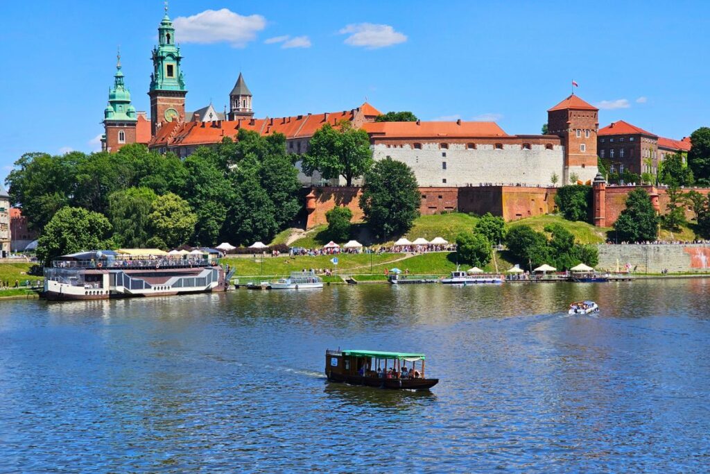 Blick über die Weichsel auf die imposante Wawel-Burg in Krakau mit ihren Türmen und Mauern, im Vordergrund fährt ein Boot auf dem Fluss.
