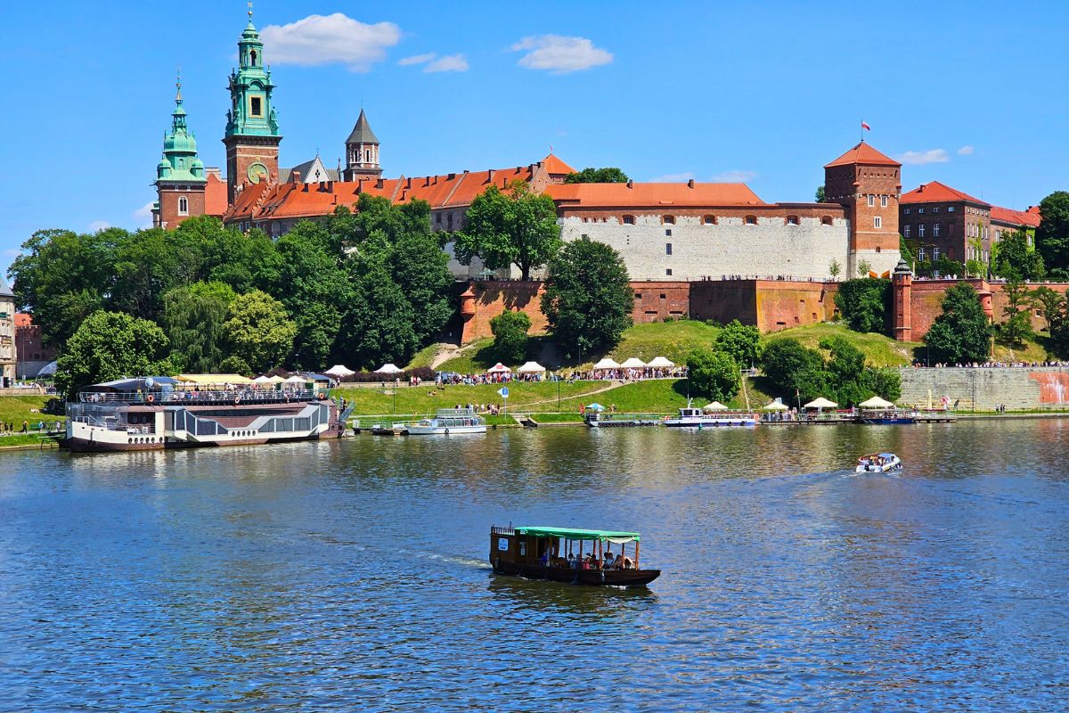 Blick über die Weichsel auf die imposante Wawel-Burg in Krakau mit ihren Türmen und Mauern, im Vordergrund fährt ein Boot auf dem Fluss.
