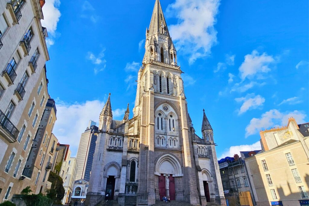 Die hohe, spitze Fassade der neugotischen Basilique Saint-Nicolas in Nantes vor einem strahlend blauen Himmel mit lockeren Wolken.