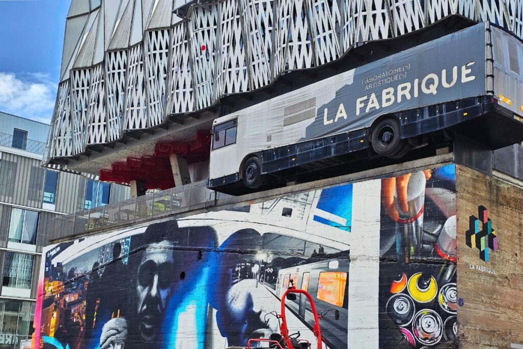 Das Gebäude „Trempolino“ in Nantes, an dessen Fassade ein echter grauer Bus zu hängen scheint, kombiniert mit bunter Streetart.