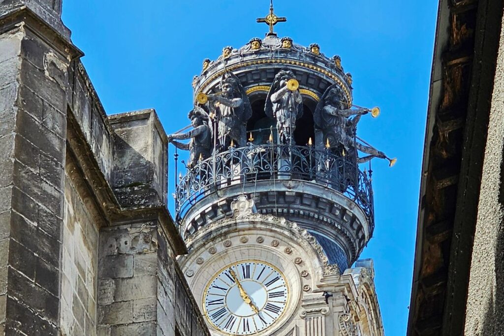 Nahaufnahme der Spitze des Uhrturms der Sainte-Croix-Kirche in Nantes mit goldenen Engeln, die Posaune blasen, vor blauem Himmel.