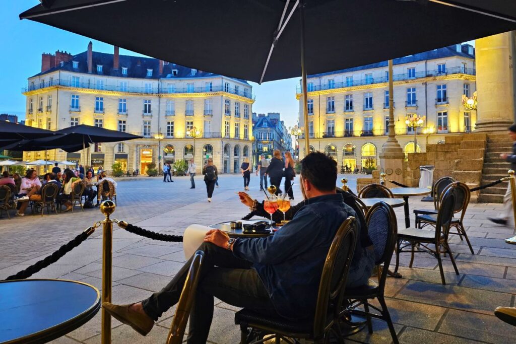 Gäste sitzen an einem Tisch im Vordergrund und genießen Drinks auf dem beleuchteten Place Graslin in Nantes während der blauen Stunde.