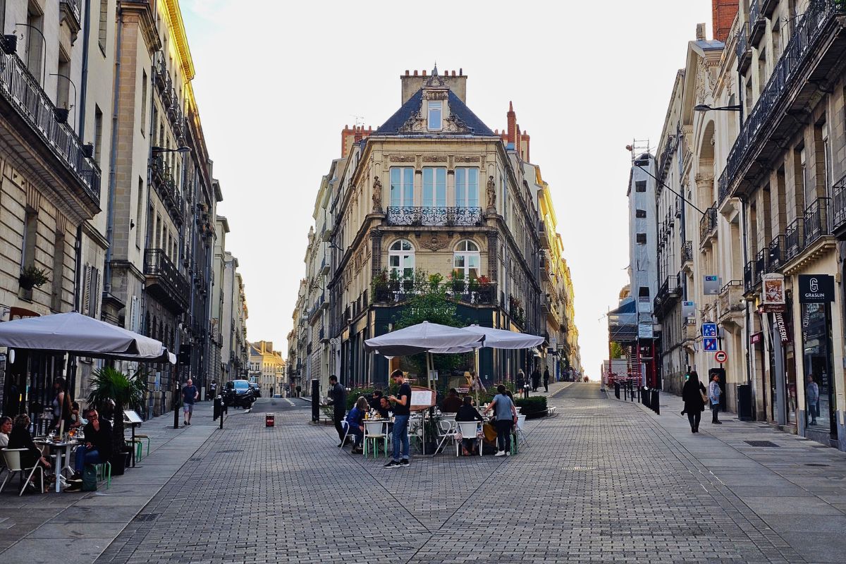 Eine weite, gepflasterte Straße in Nantes mit prachtvollen historischen Gebäuden und Menschen an Cafétischen unter strahlend hellem Himmel.