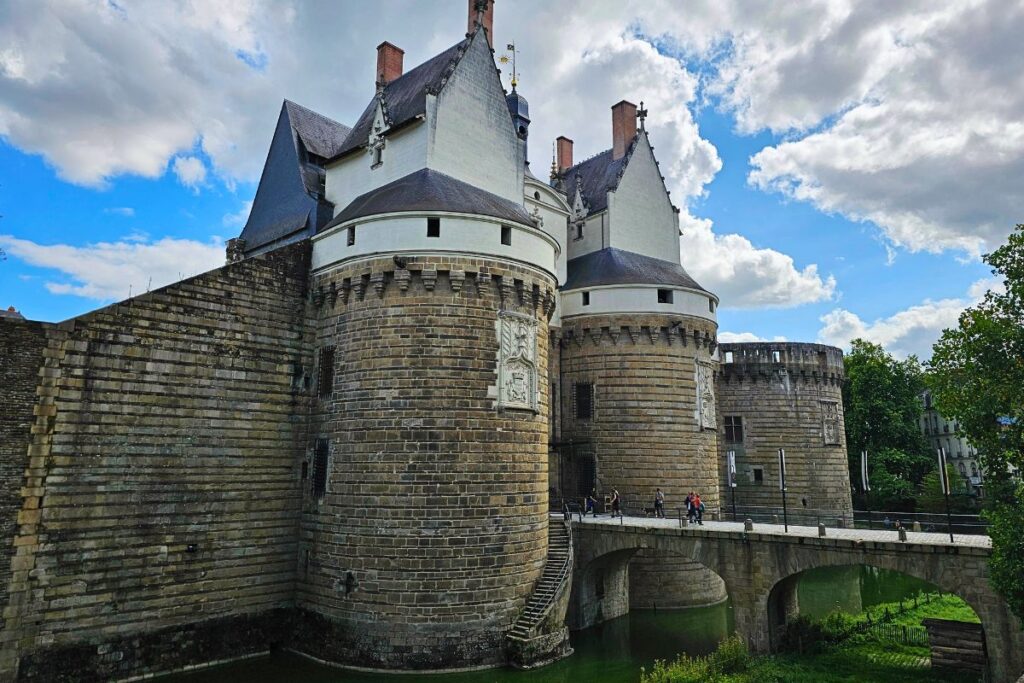 Das historische Château des Ducs de Bretagne in Nantes mit seinen massiven runden Türmen und der hellen Fassade hinter einer Steinbrücke unter bewölktem Himmel.