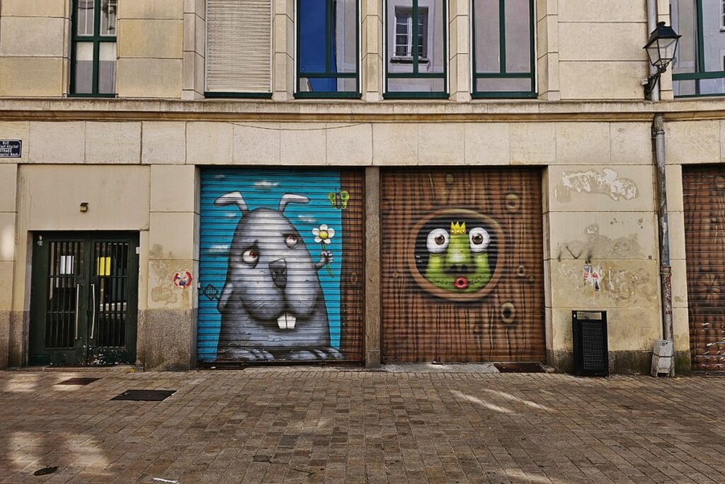 Kreative Streetart auf zwei Rollläden in Nantes; links ein grauer Hase mit einer Blume, rechts ein grüner Frosch mit Krone in einem Astloch.