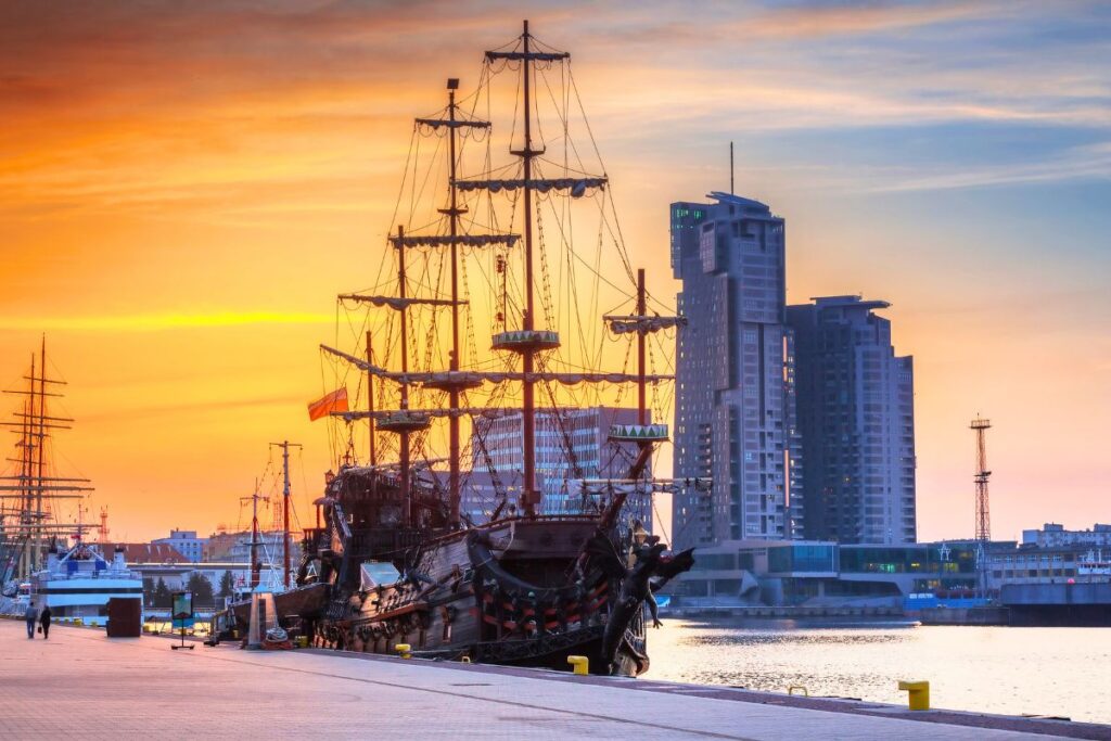 Ein hölzernes Ausflugsschiff im Piraten-Stil im Hafen von Gdingen (Gdynia) vor modernen Hochhäusern während eines goldenen Sonnenuntergangs.