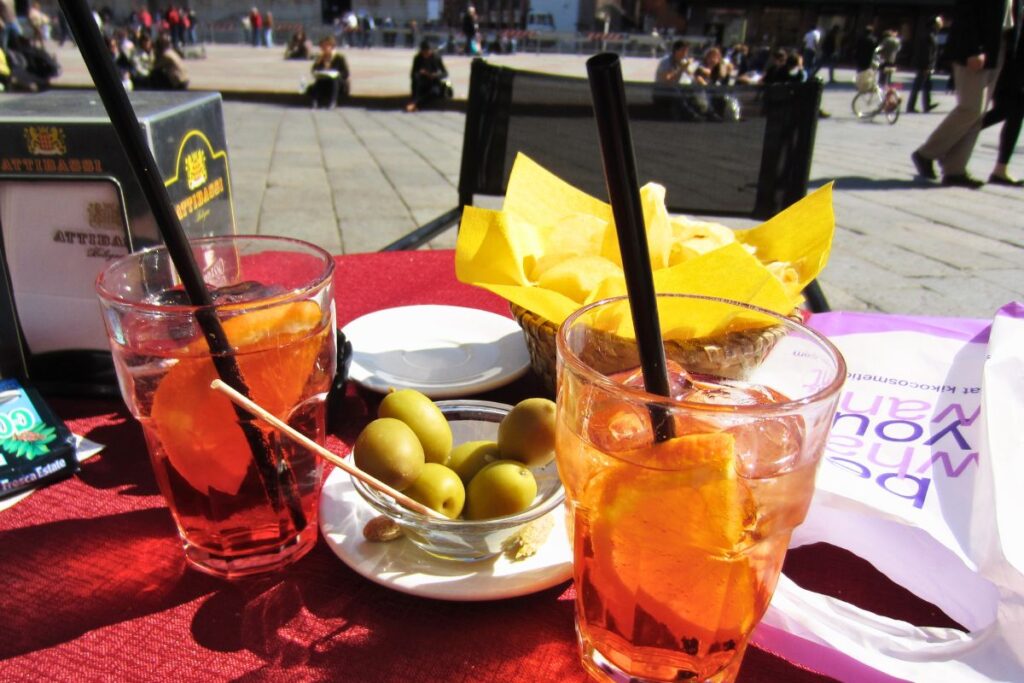 Zwei Gläser orangefarbener Spritz-Aperitif mit Oliven und Chips auf einem roten Tisch in einem sonnigen Straßencafé in Bologna.