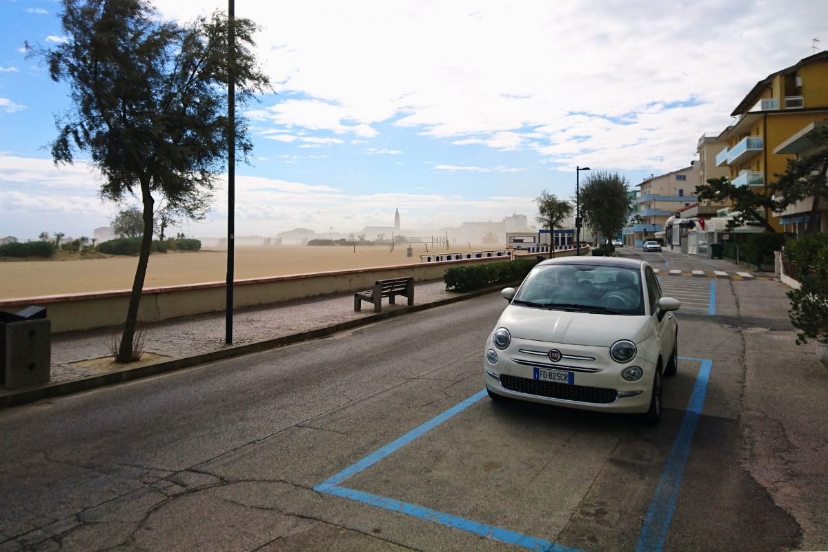 Ein kleiner weißer Fiat 500 parkt auf einer Straße direkt neben der weitläufigen Strandpromenade von Caorle.