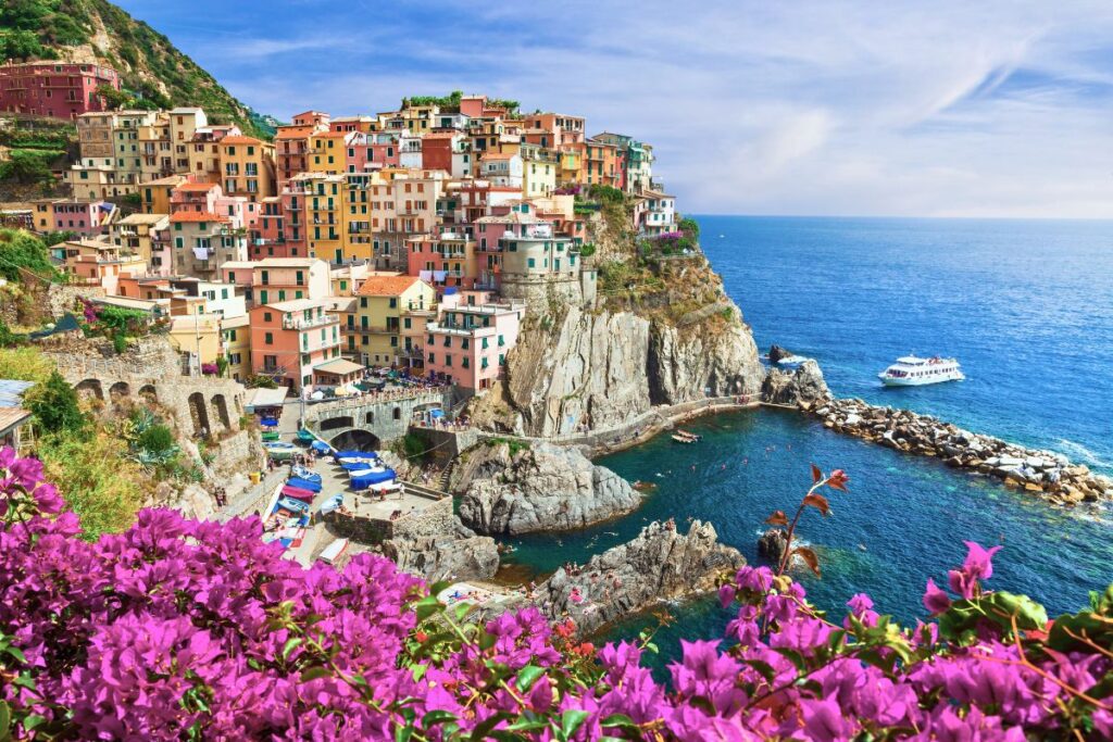 Das bunte Dorf Manarola in den Cinque Terre auf einem steilen Felsen am Meer mit leuchtend pinken Blumen im Vordergrund und einem weißen Boot.