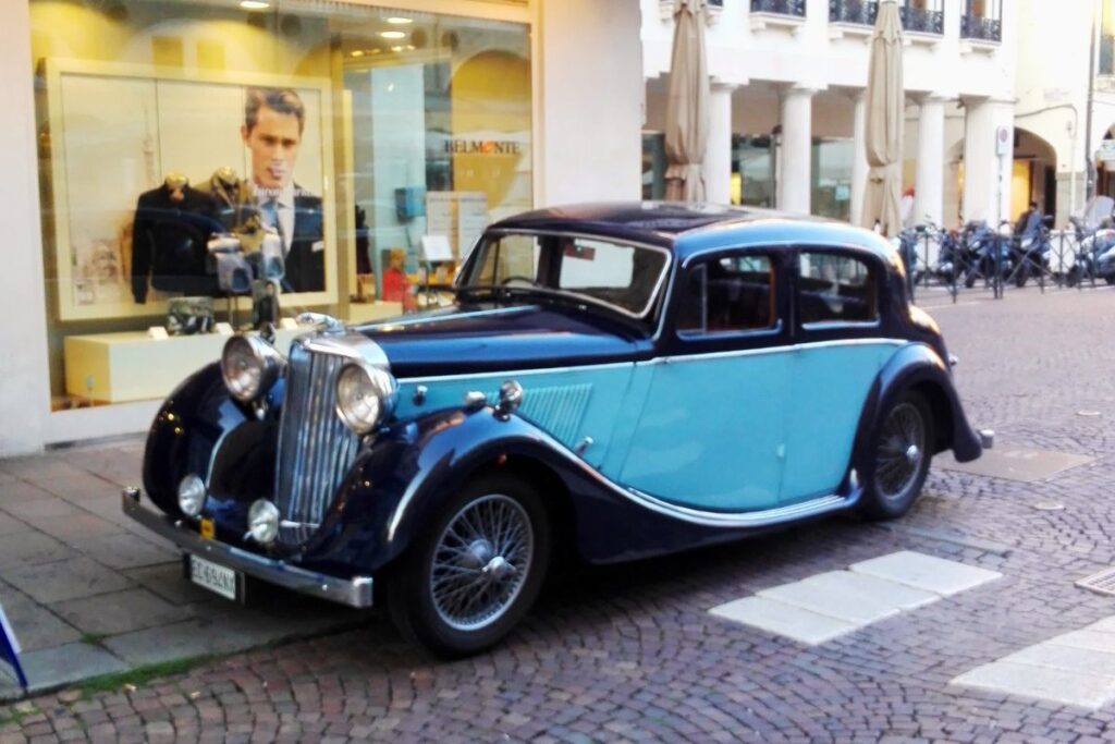 Ein stilvoller zweifarbiger Oldtimer in Dunkelblau und Hellblau parkt in einer italienischen Einkaufsstraße vor einem modernen Schaufenster.