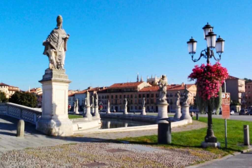 Der Prato della Valle in Padua mit seinen charakteristischen Statuen auf steinernen Podesten entlang des Kanals unter strahlend blauem Himmel.