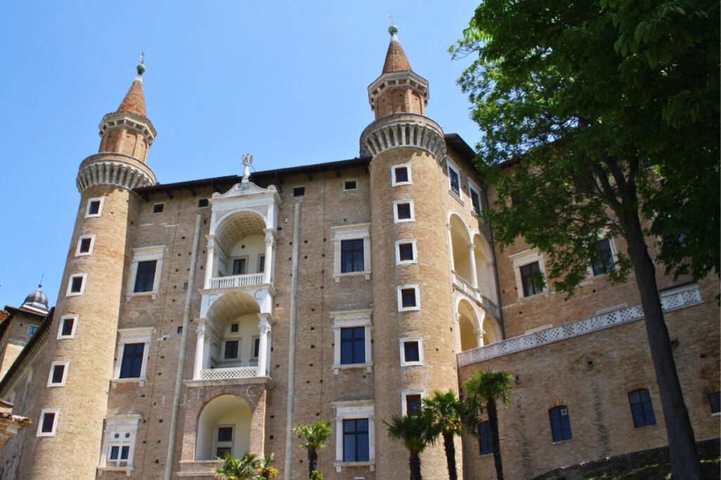 Die imposante Renaissance-Fassade des Palazzo Ducale in Urbino mit seinen markanten spitzen Zwillingstürmen und grünen Palmen im Vordergrund.