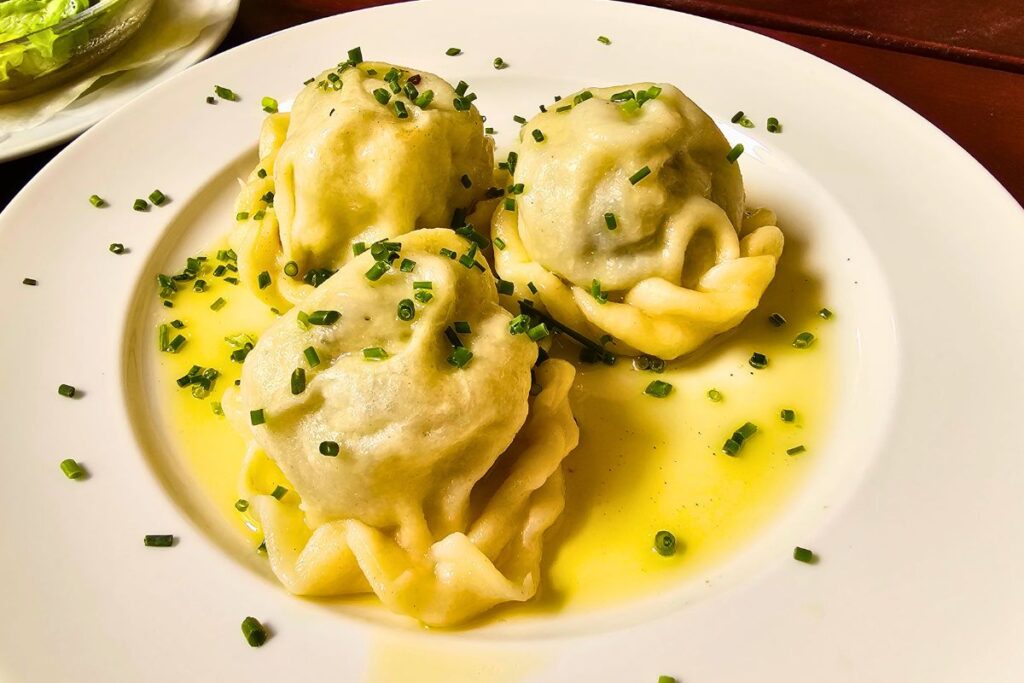 Drei traditionell gekändelte Kärntner Kasnudeln in geschmolzener Butter, garniert mit frischem Schnittlauch auf einem weißen Teller.