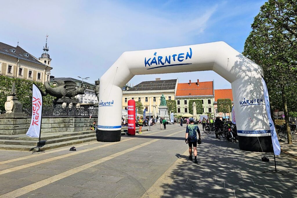 Ein Radfahrer steuert auf den großen weißen Zielbogen mit der Aufschrift „Kärnten“ auf dem Neuen Platz in Klagenfurt zu, links im Bild befindet sich der berühmte Lindwurm-Brunnen.