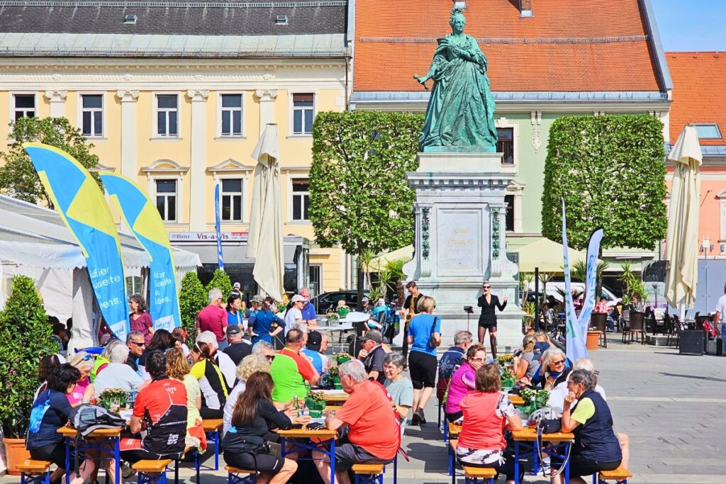 Zahlreiche Radfahrer sitzen an Bierzeltgarnituren auf dem Neuen Platz in Klagenfurt und feiern den Abschluss der Velovista-Etappe bei Live-Musik vor dem Maria-Theresia-Denkmal.