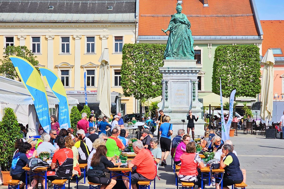 Zahlreiche Radfahrer sitzen an Bierzeltgarnituren auf dem Neuen Platz in Klagenfurt und feiern den Abschluss der Velovista-Etappe bei Live-Musik vor dem Maria-Theresia-Denkmal.